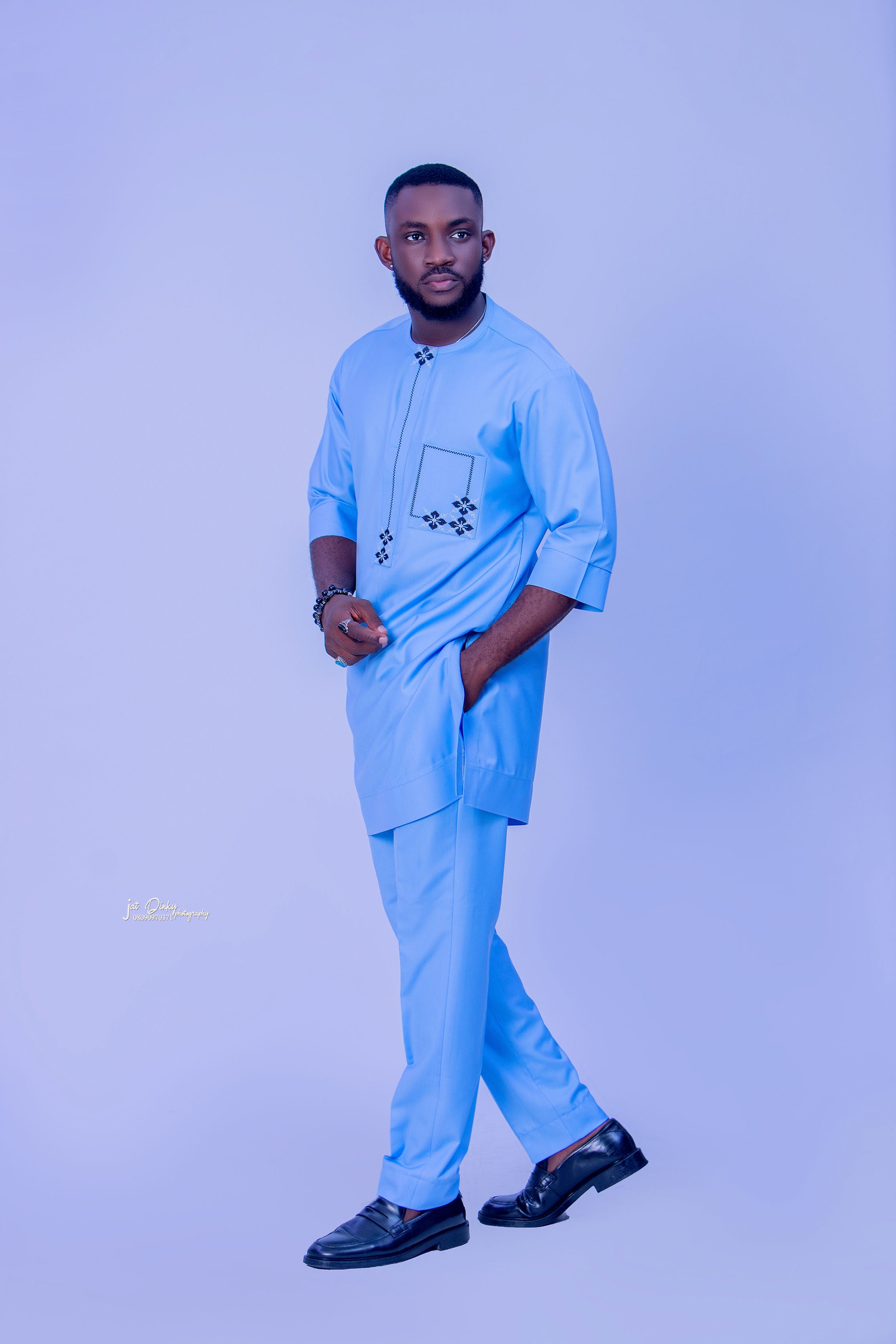 A man wearing a traditional African blue kaftan with intricate detailing on the neckline and pocket, standing against a purple background. Ready to wear & Luxurious.